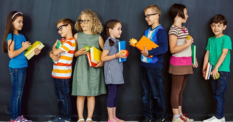 Seven young school children talking with each other, boys and girls, each holding books and an apple