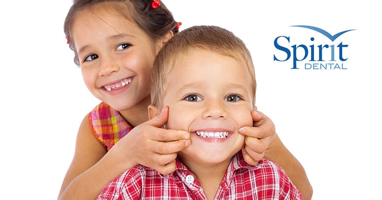 A young girl sitting behind a young boy, smiling as she holds his cheeks in a smiling position.  Both have very white teeth.  